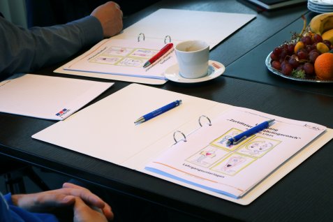Mape cu documente de instruire și pixuri pe o masă la care sunt așezați angajații.